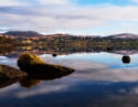 Calm lake reflecting mountains and clouds with rocks in the foreground.