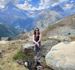 Person sitting on a rock with a dog, mountainous backdrop with clear skies.