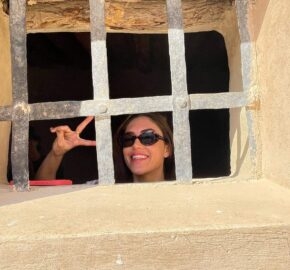 A smiling woman making a peace sign behind a barred window in a stone wall.