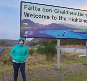 A person in a teal jacket stands in front of a 'Welcome to the Highlands' sign with a scenic backdrop.