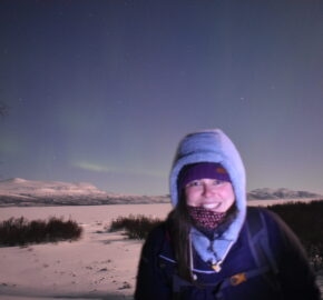 Person in winter clothing with snowy landscape and faint aurora in starry night sky.