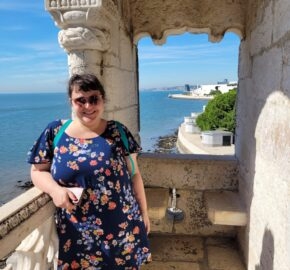 Person in floral dress standing in stone archway overlooking sea and coastline.