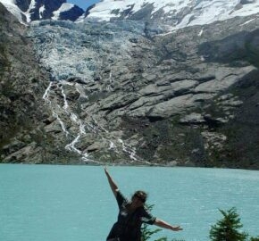 Person posing with arms outstretched on a rock, with a glacier and turquoise lake in the background.
