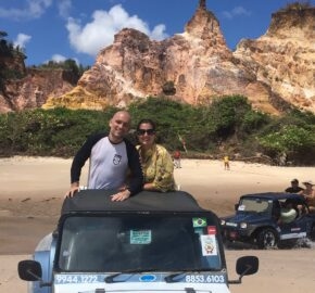 Two people leaning on a jeep with colorful cliffs in the background on a sunny beach day.