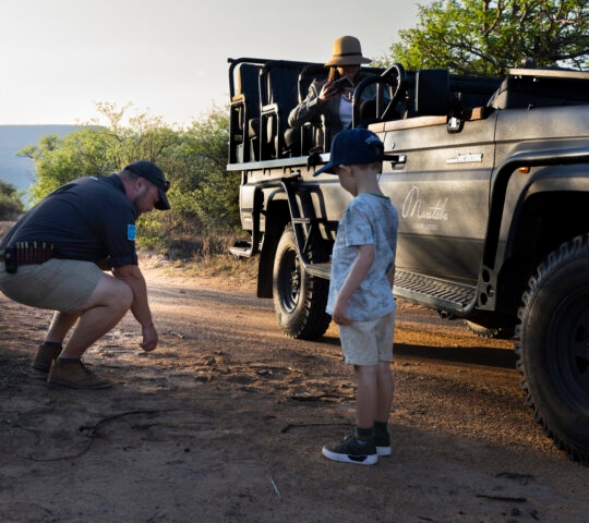 An adult crouching and a child standing near a safari vehicle with passengers in a dusty terrain.