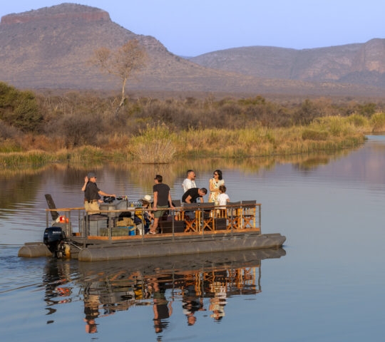 A group of people on a floating deck with a table on a calm river, with a mountainous backdrop.