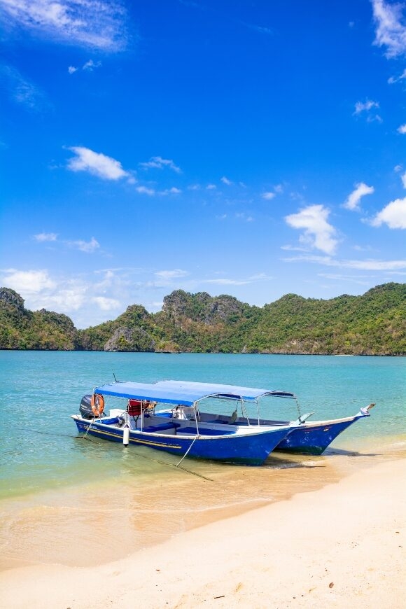 A blue and white boat moored on a sunny tropical beach with clear water and green hills.