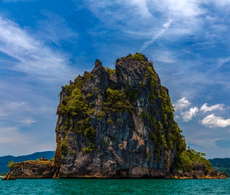 Rocky island with lush greenery in turquoise sea under blue sky with clouds.