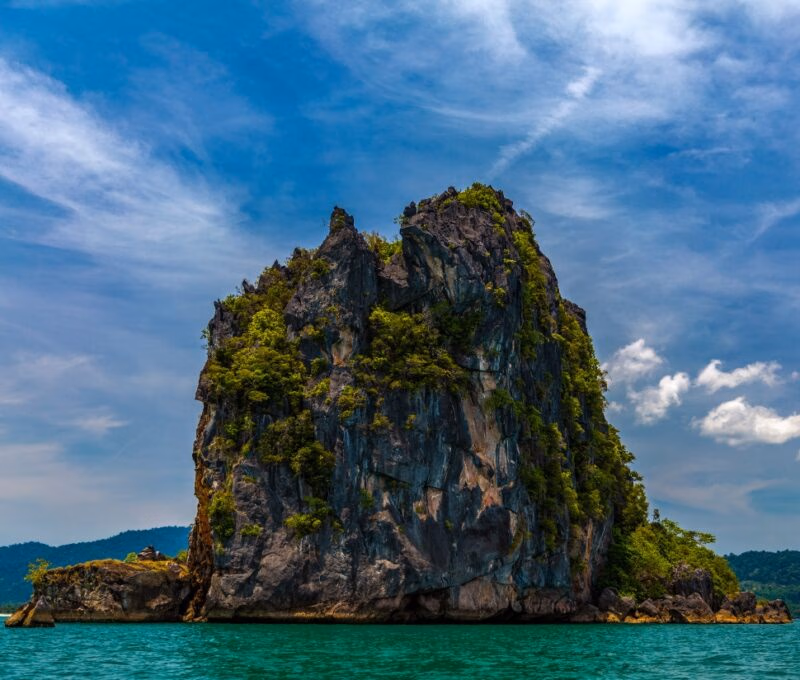 Rocky island with lush greenery in turquoise sea under blue sky with clouds.