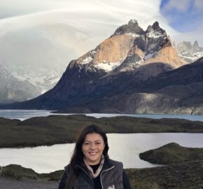 Person in a jacket standing before a scenic mountain and lake landscape.