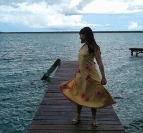 Woman in a yellow dress standing on a wooden dock over a body of water.
