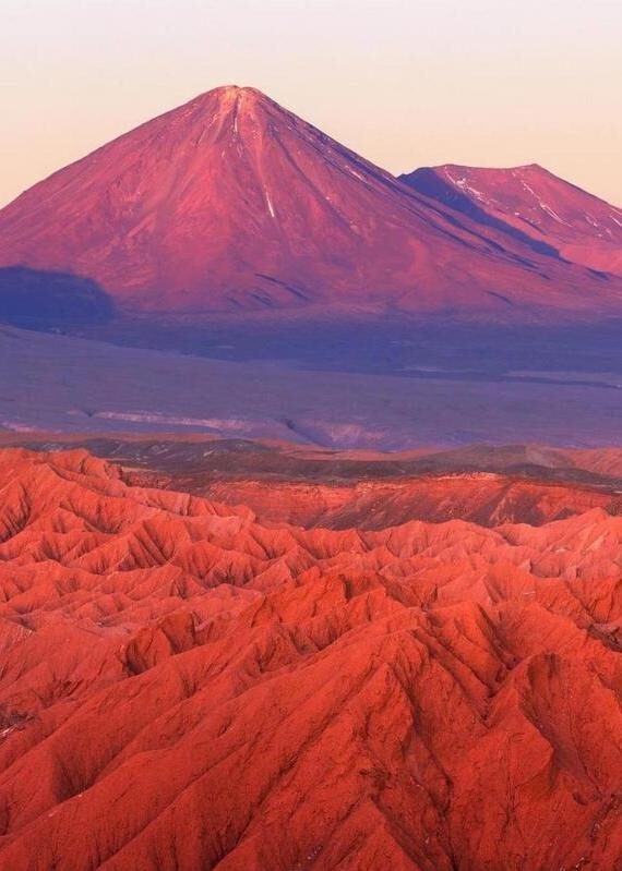 Reddish hues bathe a rugged landscape with a conical volcano in the background.
