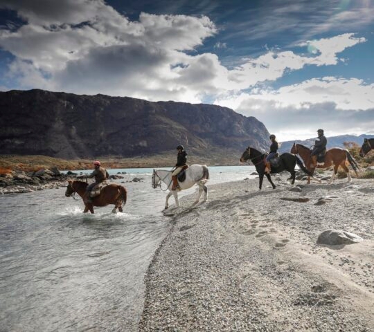 Riders on horses crossing a shallow river with a mountainous backdrop under a cloudy sky.