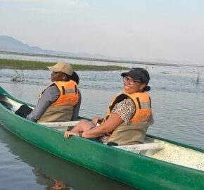 Two individuals in life jackets sitting in a green canoe on calm water with mountains in the background.