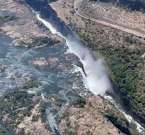 Aerial view of a waterfall with mist, surrounded by a rugged landscape and a bridge.
