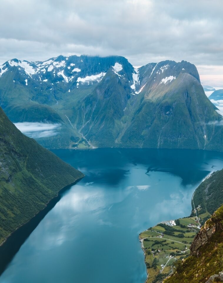 Aerial view of a fjord with a village amidst mountains and snow patches.