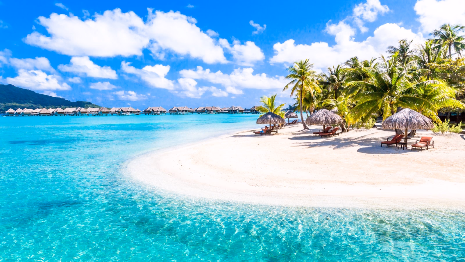 Tropical beach with clear blue water, white sand, palm trees, thatched umbrellas, and overwater bungalows.