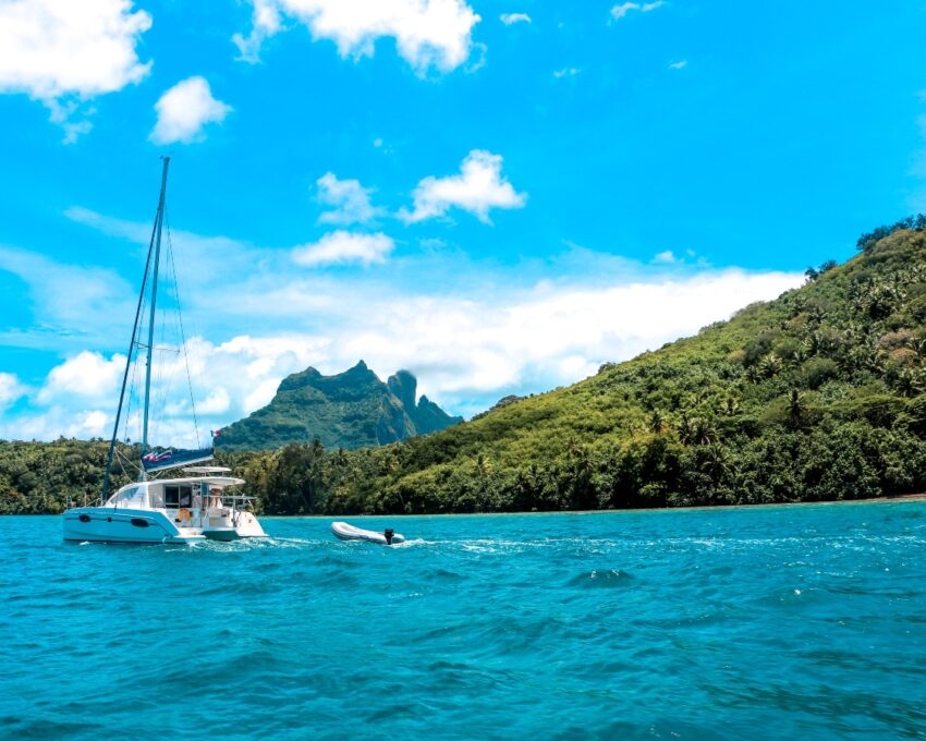 Catamaran and dinghy on blue water with tropical green mountainous background under a blue sky.