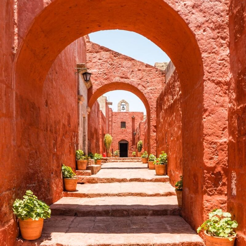 Luxury Peru Tours - Arched pathway in a red-walled monastery with potted plants under bright sunlight.