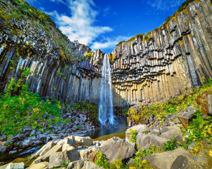 Waterfall cascading over a unique cliff of columnar basalt under a blue sky.
