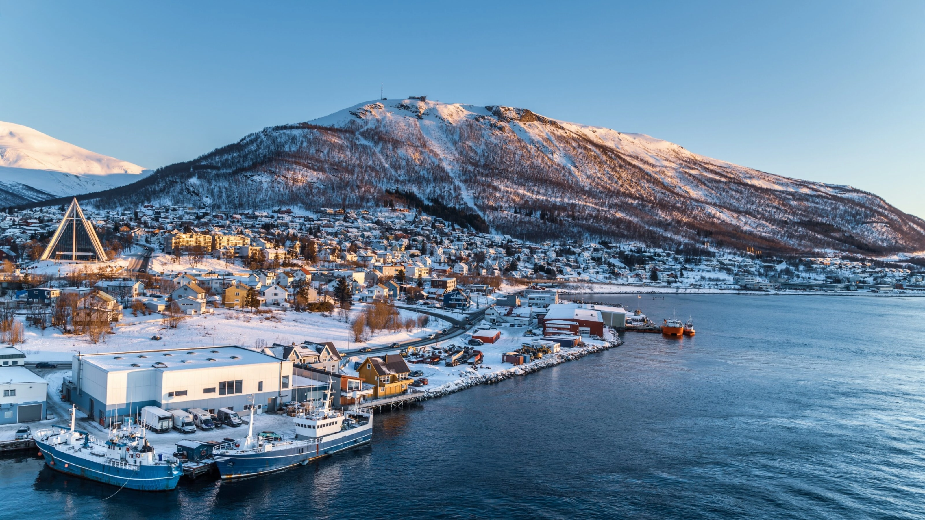 Snowy coastal town with boats and a triangular church during sunset.