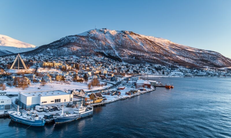 Snowy coastal town with boats and a triangular church during sunset.