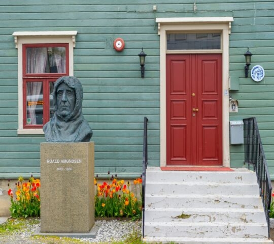A statue of Roald Amundsen in front of a green house with a red door and blooming tulips.