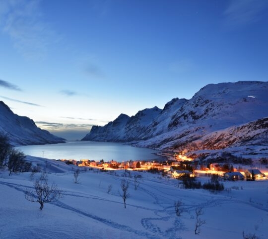 Twilight over a snow-covered village by a fjord, with mountains under a starry sky.