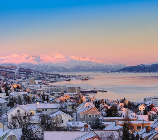 Winter sunrise over a snow-covered coastal town with mountains in the background.