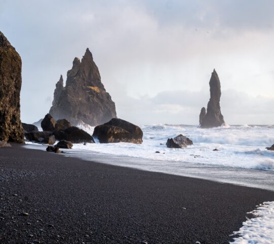 A black sand beach with jagged rocks and sea stacks under a cloudy sky.