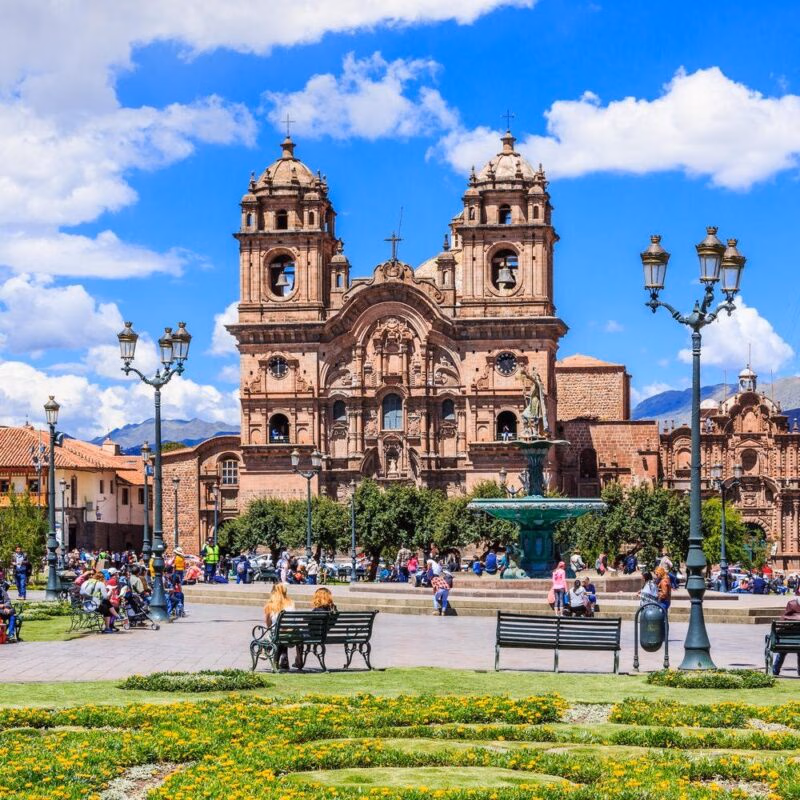 Colonial cathedral at a lively plaza with pedestrians, benches, and greenery under a blue sky.