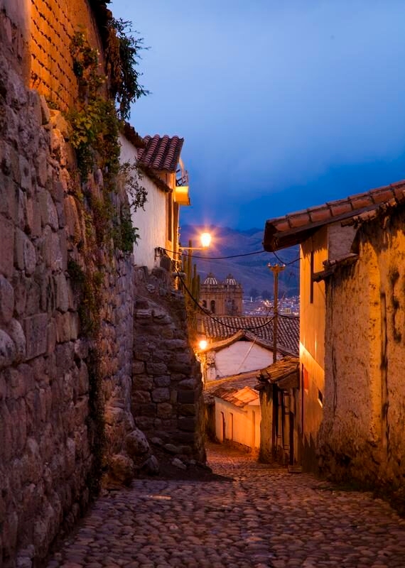 Narrow cobblestone street at twilight with stone walls and a distant church view.
