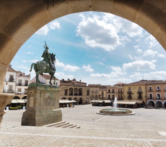 Equestrian statue in a plaza with a fountain and surrounding buildings, viewed through an archway under a blue sky.