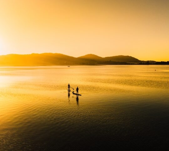 Two people paddleboarding on calm water at sunset with mountains in the background.