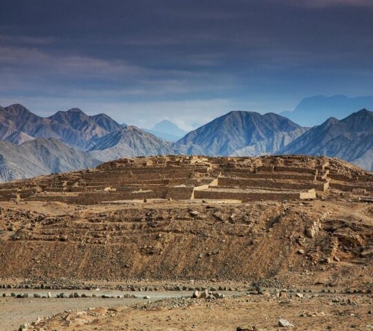 Layered ancient ruins with a backdrop of rugged mountains under a blue sky.
