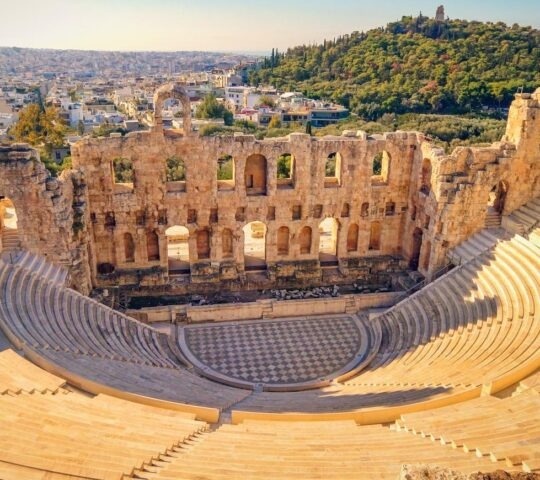 Aerial view of an ancient amphitheater ruins with cityscape and forested hill in the background.