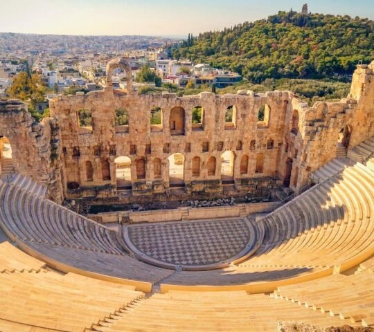 Aerial view of an ancient amphitheater ruins with cityscape and forested hill in the background.