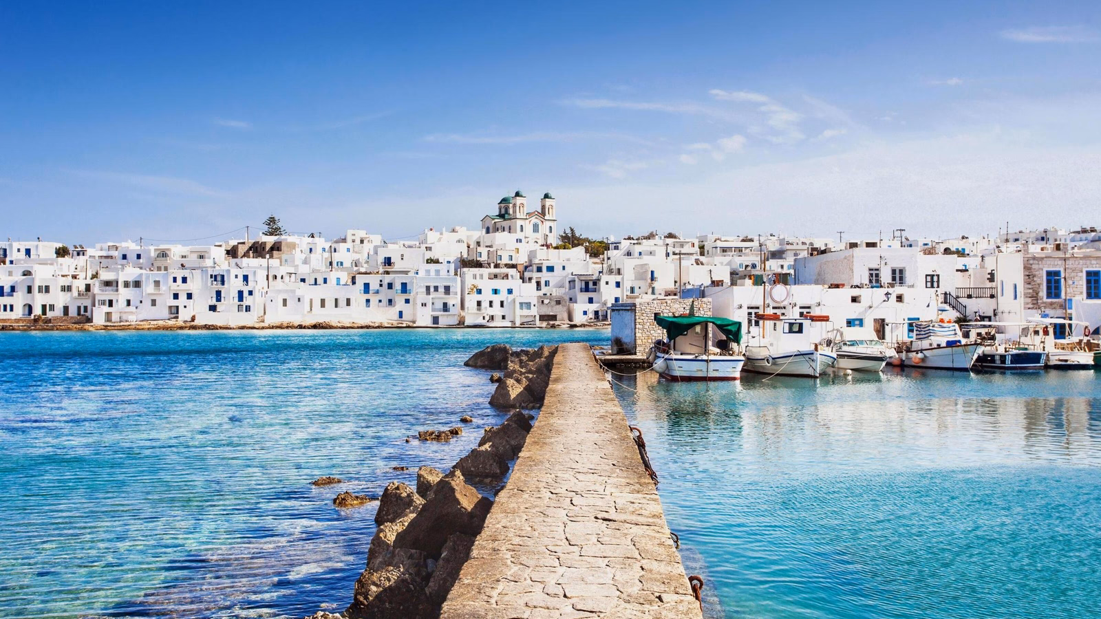 White buildings with blue domes in a Greek island town by the sea with moored boats.