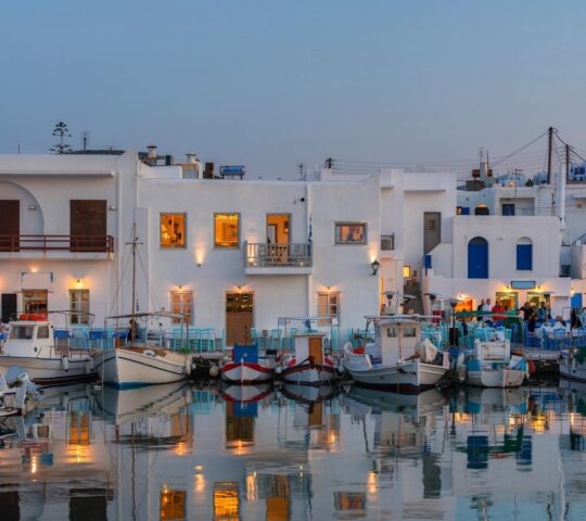Twilight at a serene Mediterranean harbor with boats and adjacent dining area.