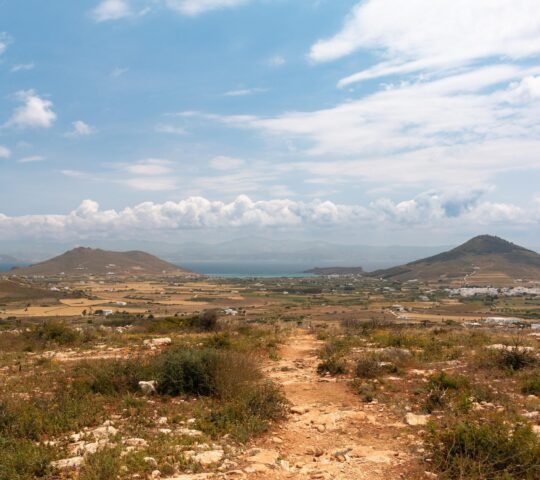 Rural landscape with rolling hills, fields, and a dirt path under a blue cloudy sky.