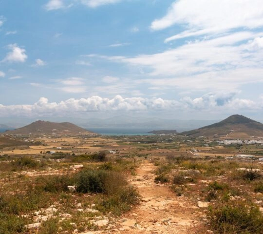 Rural landscape with rolling hills, fields, and a dirt path under a blue cloudy sky.