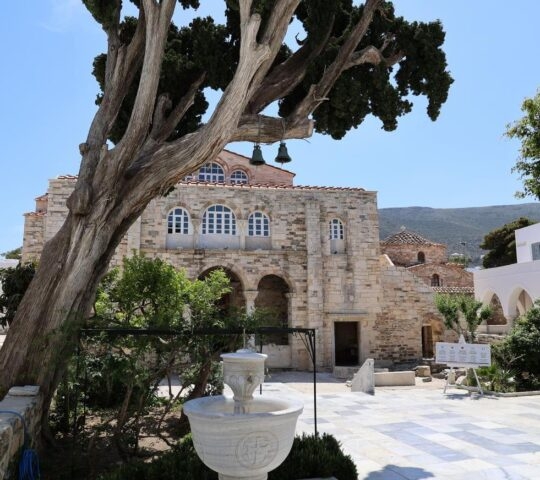 Ancient stone church with arched entrance, flanked by trees and a fountain in the courtyard.