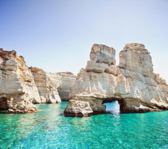 Rocky cliffs with a natural arch over clear turquoise sea water under a blue sky.