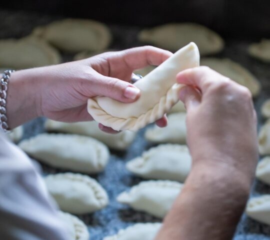 Hands crimping the edge of a pastry on a table with more pastries in the background.
