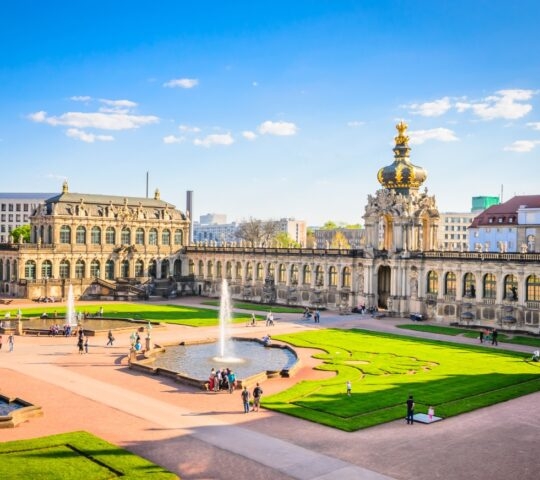 Baroque palace with fountain, ornate architecture, and visitors enjoying the gardens under a clear sky.