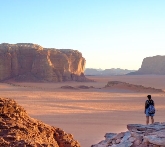 Person standing on rock overlooking vast desert and towering cliffs at sunset.