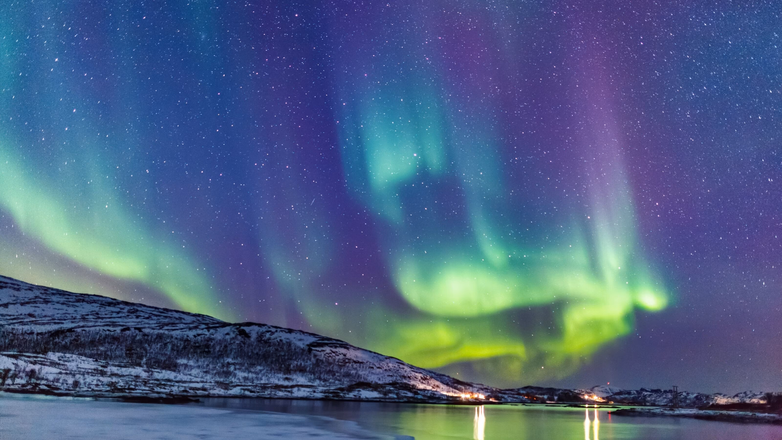 A vivid aurora borealis above a snowy landscape and icy water with stars twinkling in the sky.