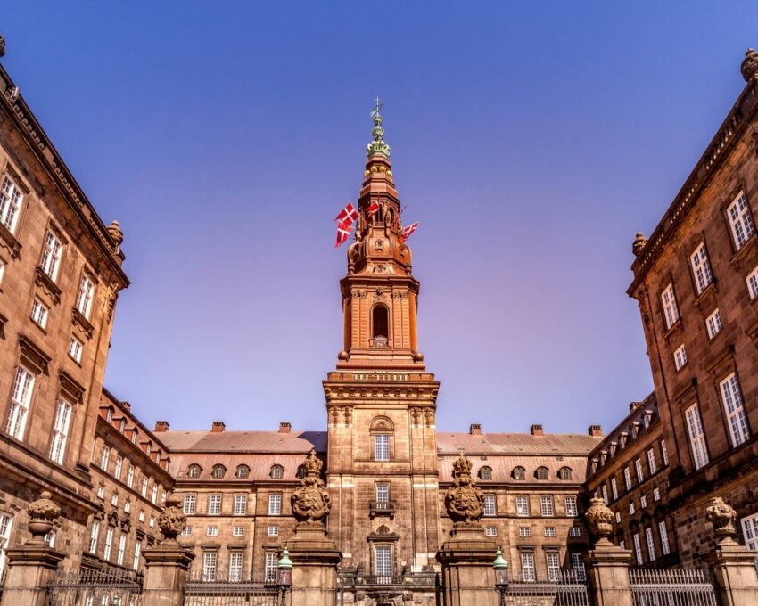 Facade of a historic European palace with a spire and Danish flag under a clear blue sky