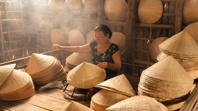 A person works among stacks of traditional conical hats in a workshop.