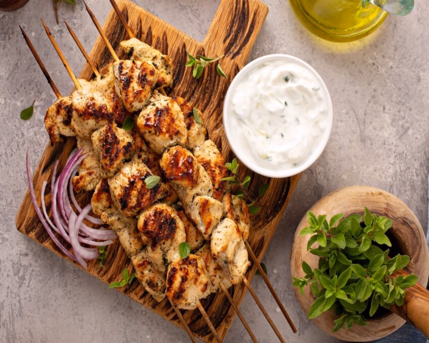 Grilled chicken skewers on a wooden board with tzatziki sauce and fresh herbs.